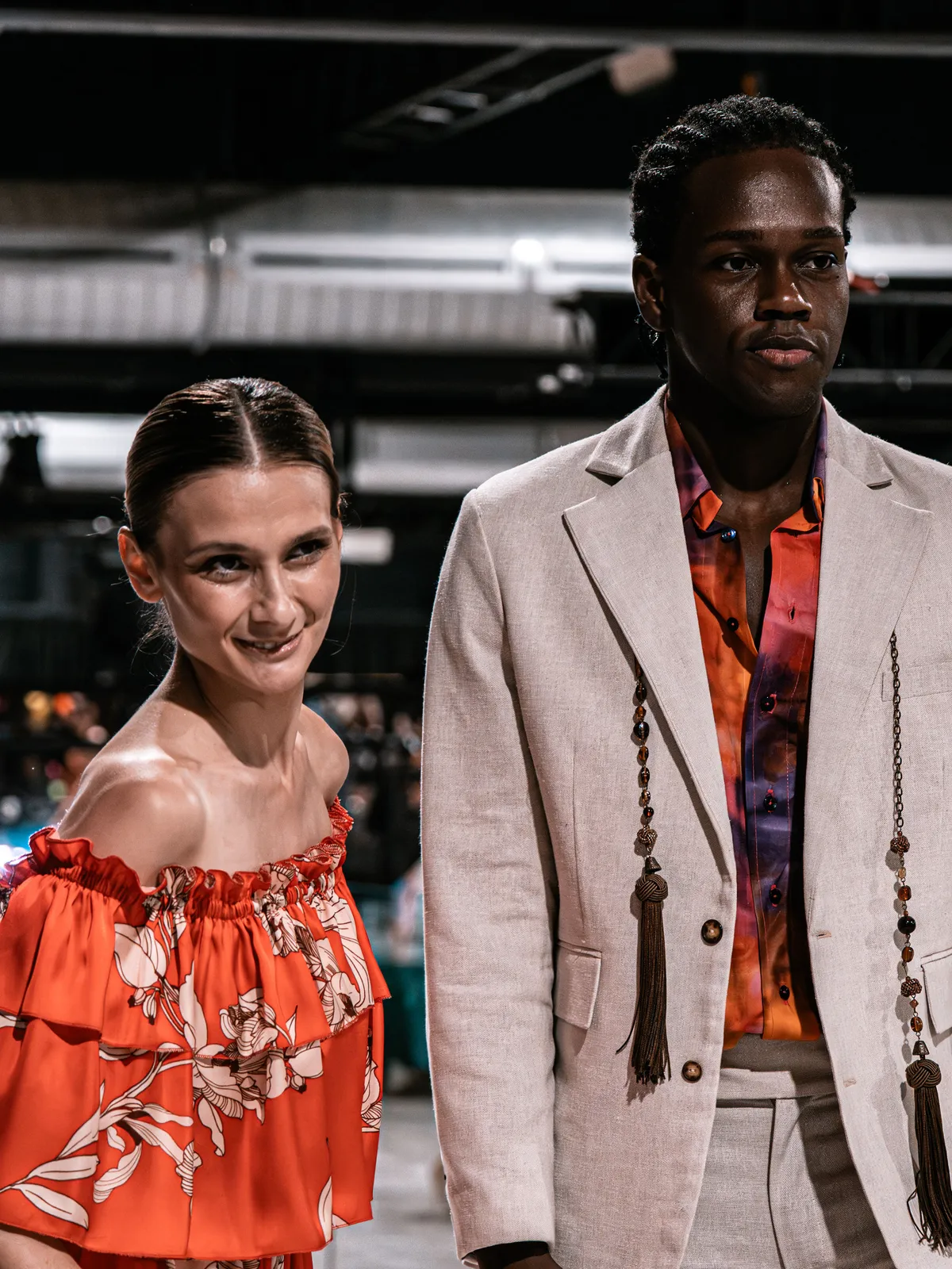 Dos models al final de la passarel·la del 2025: ella amb un vestit taronja de flors i ell amb un vestit de lli clar