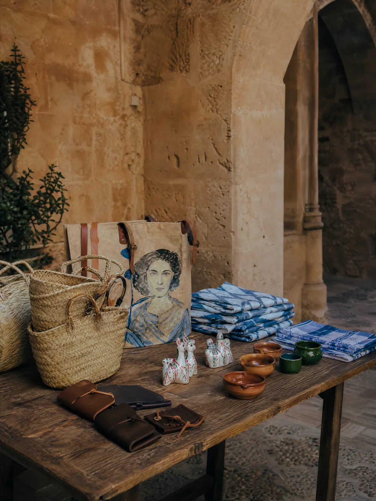 Palmblattkörbe, eine mit einem Porträt bemalte Jutetasche, blauer Llengües-Stoff, Siurells und Keramikschalen auf einem Holztisch, in einem Innenhof aus Marès-Stein